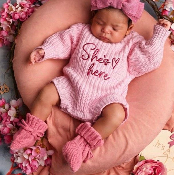 Newborn baby in a pink 'She's here' outfit with dusky pink bow and booties