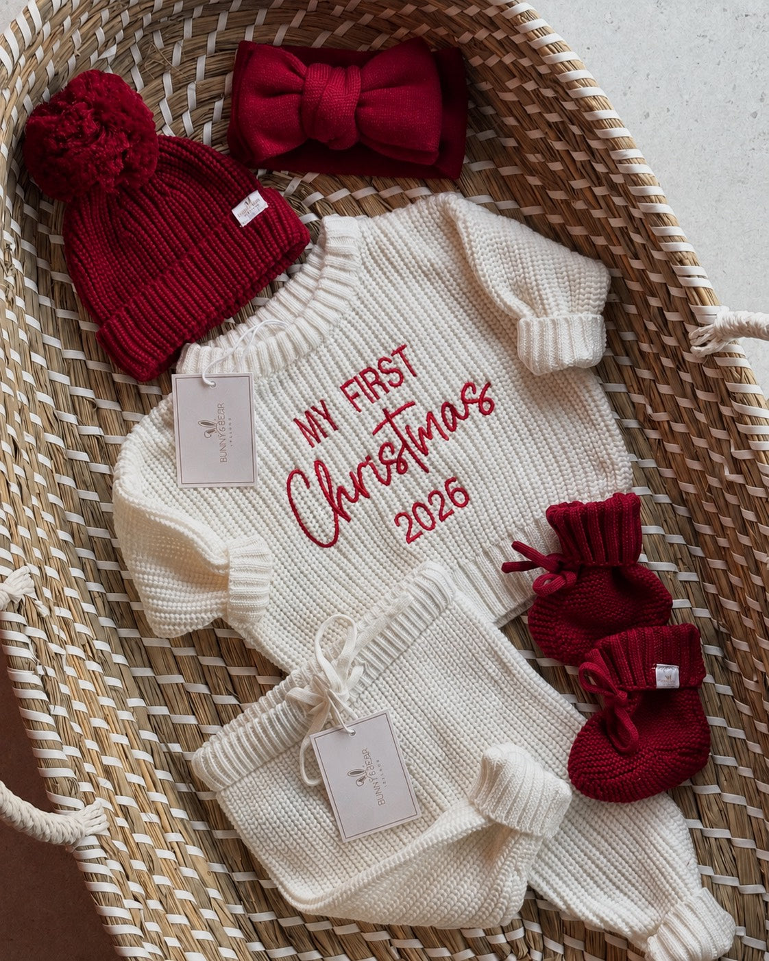 A baby knit holiday outfit set arranged in a woven basket, including a cream ribbed long-sleeve romper with red embroidered text reading 'My First Christmas 2026,' a matching cream knit hat, cream booties, and dark red knit accessories such as a bow, pom-pom hat, and mittens.